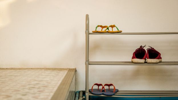 Minimal Metal Shoe Rack with Two Pairs of Flip Flops and a Pair of Red Shoes. White Wall Texture. Blue/ Teal Water Pipe.