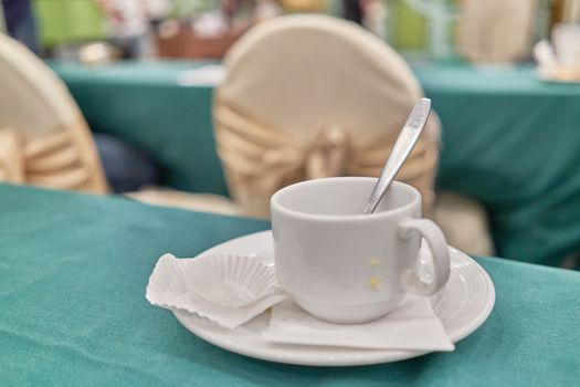 Empty unclean white cup of coffee with spoon and tissue with empty paper cup of cookie and cake on saucer are placed on green table have blur gold chair of conference in the hotel as background.