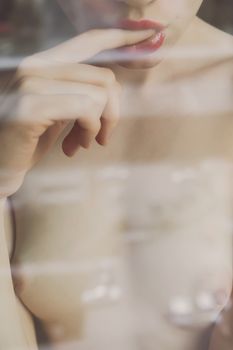 Crop view through glass of faceless naked young woman sexually putting finger in mouth