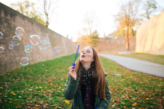 Attractive young woman is enjoying making a bubbles in the public park. Ground covered with autumn leaves.