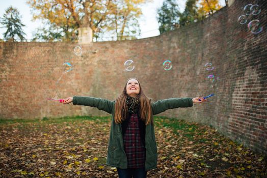 Attractive young woman is enjoying making a bubbles in the public park with raised hands. Ground covered with autumn leaves.