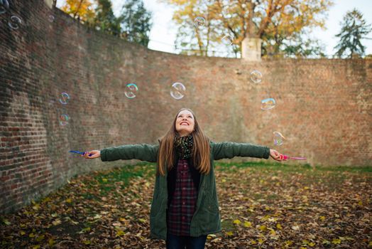 Attractive young woman is enjoying making a bubbles in the public park with raised hands. Ground covered with autumn leaves.