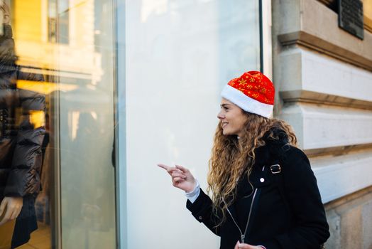 Young attractive woman with santa claus hat in front of shopping window looking to buy something