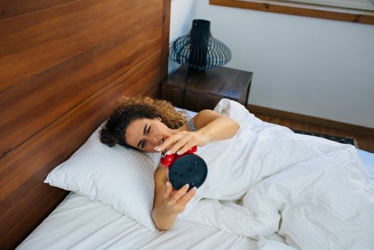 Beautiful woman awakened by alarm clock in the bed at morning time. A woman is unhappy from noisy sound of alarm clock and trying to turn it off.