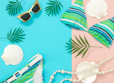 Summer fashion flat lay on blue and pink background. Sunglasses, bikini, pearl necklace, seashells and palm leaves.
