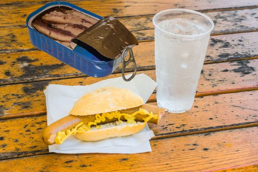 Bockwurst in bun, a can of fish, mug with mineral water on a brown wooden table on a white napkin.