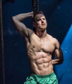 Attractive shirtless blond male bodybuilder in shorts indoors in dark gym, showing muscular torso and ripped abs