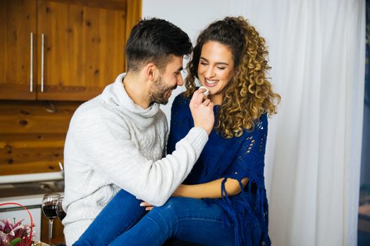 Young handsome man and his attractive seductive girlfriend is having sweet emotions in the kitchen with cookies and wine. Man is giving a cookie to his love.