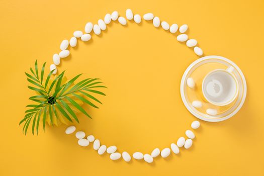 Frame made of white chocolate candies, palm tree leaves and teacup, on bright yellow background.