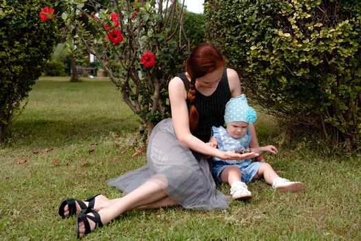 The girl shows the child the bumps in the palm, the baby looks with interest. They sit in the Park on the grass, against a Bush with red flowers