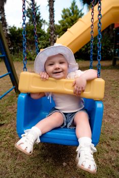 the baby is smiling from a fun trip on a swing. She plays in a blue swing, on the street, in the Playground, wearing a hat