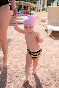 the girl goes hand in hand with her mother on the large sand on a public beach, between the beach loungers, she is dressed in a pink cap and striped swimming panties