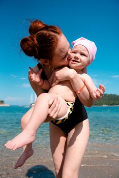 girl playing with a child standing on the beach near the water. they're both wearing bathing suits, blue sky and sea in the back.