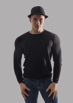 Handsome young muscular man looking at camera in studio shot, wearing fedora hat, with arms crossed on chest, on grey background