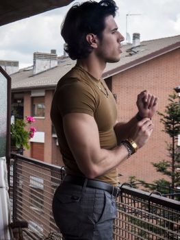 Handsome dark haired young man standing in a balcony, looking out and down at the view, wearing brown T-shirt, in summer