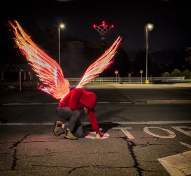 Fallen male angel kneeling in city parking lot, with fire wings spreading from his back. Unrecognizable young man