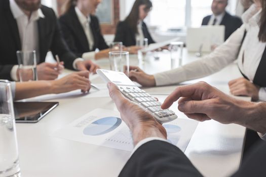 Group of business people in meeting room at office discuss financial reports, focus on male hands counting on calculator