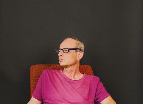 Creative portrait. Middle aged man with glasses sitting in an armchair. Portrait of mature man on black background. Copy space.