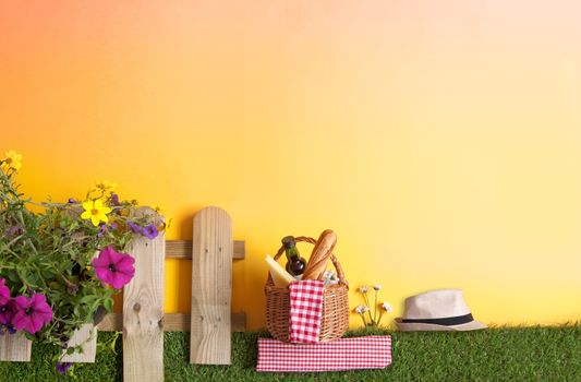 Summer picnic basket with bread, cheese and wine on grass with garden fence and flowers