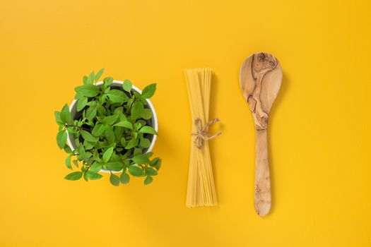 Cooking pasta. Basil herbs in a pot, spaghetti and wooden spoon on yellow background.