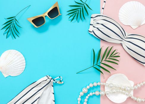 Tropical summer vacation flat lay on blue and pink background, with copy space. Bikini, sunglasses, palm leaves, seashells and pearl necklace.