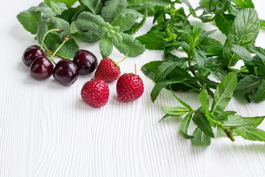 Vinous cherries, red strawberries with green herbal mix of fresh mint and melissa herbs on white wooden background