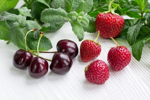 Vinous cherries, red strawberries with green herbal mix of fresh mint and melissa herbs on white wooden background