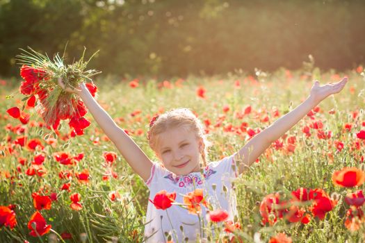 Young girl with bouquet among poppies field at sunset with hands up