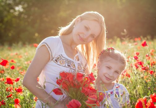 Mother brunette in a white dress with daughter together kissing on blossoming red poppies field