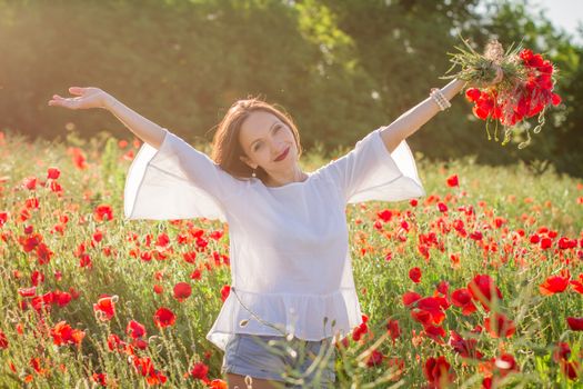 Woman with bouquet among poppies field at sunset with hands up