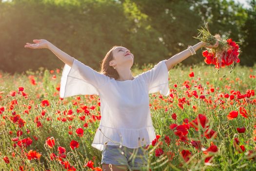 Woman with bouquet among poppies field at sunset with hands up