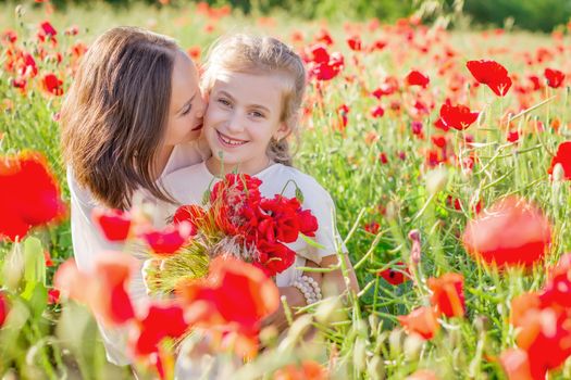 Mother brunette in a white dress with daughter together kissing on blossoming red poppies field