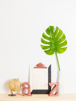 Modern home decor. Blank sheet of paper with copy space on a clipboard, green leaf, globe and metal letters on a wooden shelf.
