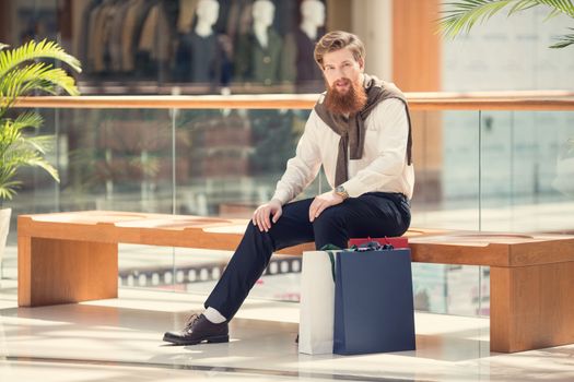 Sale and people - young bearded man with shopping bags sitting in shopping mall