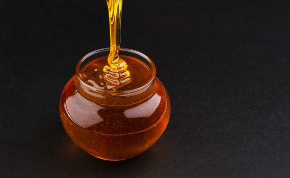 Glass pot of pouring honey isolated on black background with copy space