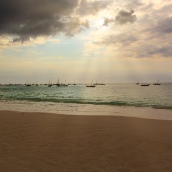 Several fishing boats silhouette anchored out in the ocean off the coast of Zanzibar, at sunset with sunbeams.