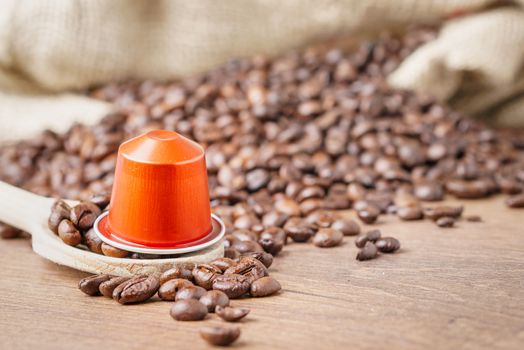 In the foreground a coffee capsule on wooden spoon and   roasted coffee beans with burlap sack on blur wooden background ,close up.