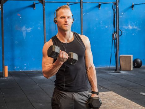 Handsome blond young man training biceps lifting dumbbells, standing in a gym, looking to a side
