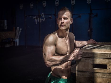 Attractive shirtless blond male bodybuilder in shorts indoors in dark gym, showing muscular torso and ripped abs