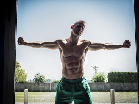 Attractive shirtless blond male bodybuilder in shorts indoors against door glass, showing muscular torso and ripped abs