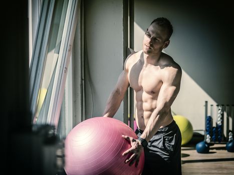 Attractive blond young man shirtless in gym working out, holding inflatable fitness ball, about to do exercises for abs.