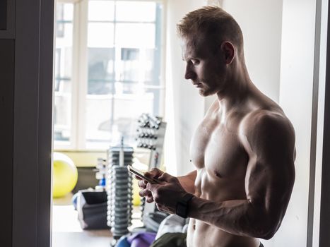 Shirtless muscular blond young man using cell phone in gym