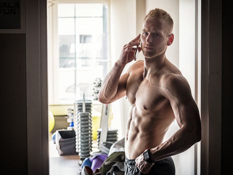 Shirtless muscular blond young man using cell phone in gym, calling somebody