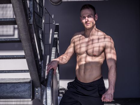 Portrait of a sexy muscular young man shirtless in urban environment, walking on metal stairs, looking at camera