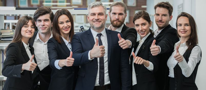 Group of friendly businesspeople with male mature leader in office showing thumbs up sign