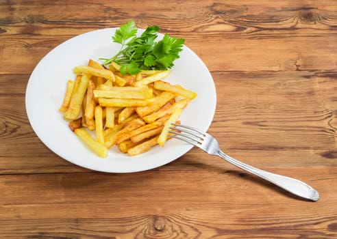 French fries with several twigs of parsley on the white dish and fork of stainless steel on a surface of old wooden planks 
