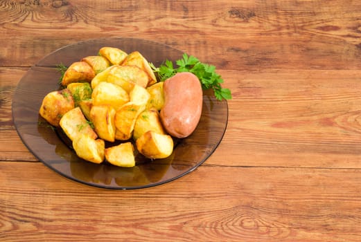 Serving of the country style fried potatoes sprinkled by chopped dill, fried wieners and twig of parsley on dark glass dish on a surface of old wooden planks
