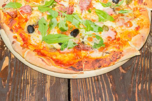 Top view of a fragment of the cooked round pizza with sausages, mushrooms, olives and arugula on the old wooden table closeup
