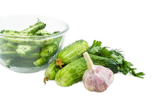 Fragment of the glass bowl with lightly salted cucumbers and several freshly picked out cucumbers, parsley, dill and garlic beside on a white background
