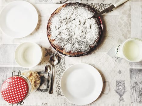 Aerial view of a table set for breakfast with white plates, teaspoons and a cake with powdered sugar